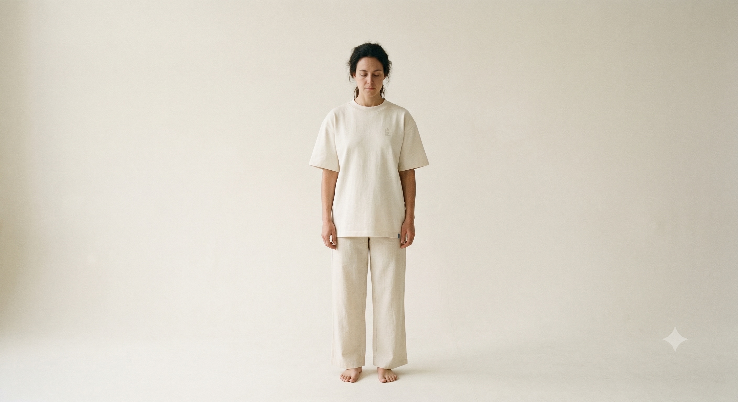 A person stands square to camera in soft north-light against a seamless ecru studio backdrop, wearing a boxed heavyweight ecru cotton crew T-shirt with dropped shoulders and one even hem; a tiny tonal (È) seal sits at the left chest and a single ink-blue tack stitch marks the left side hem.