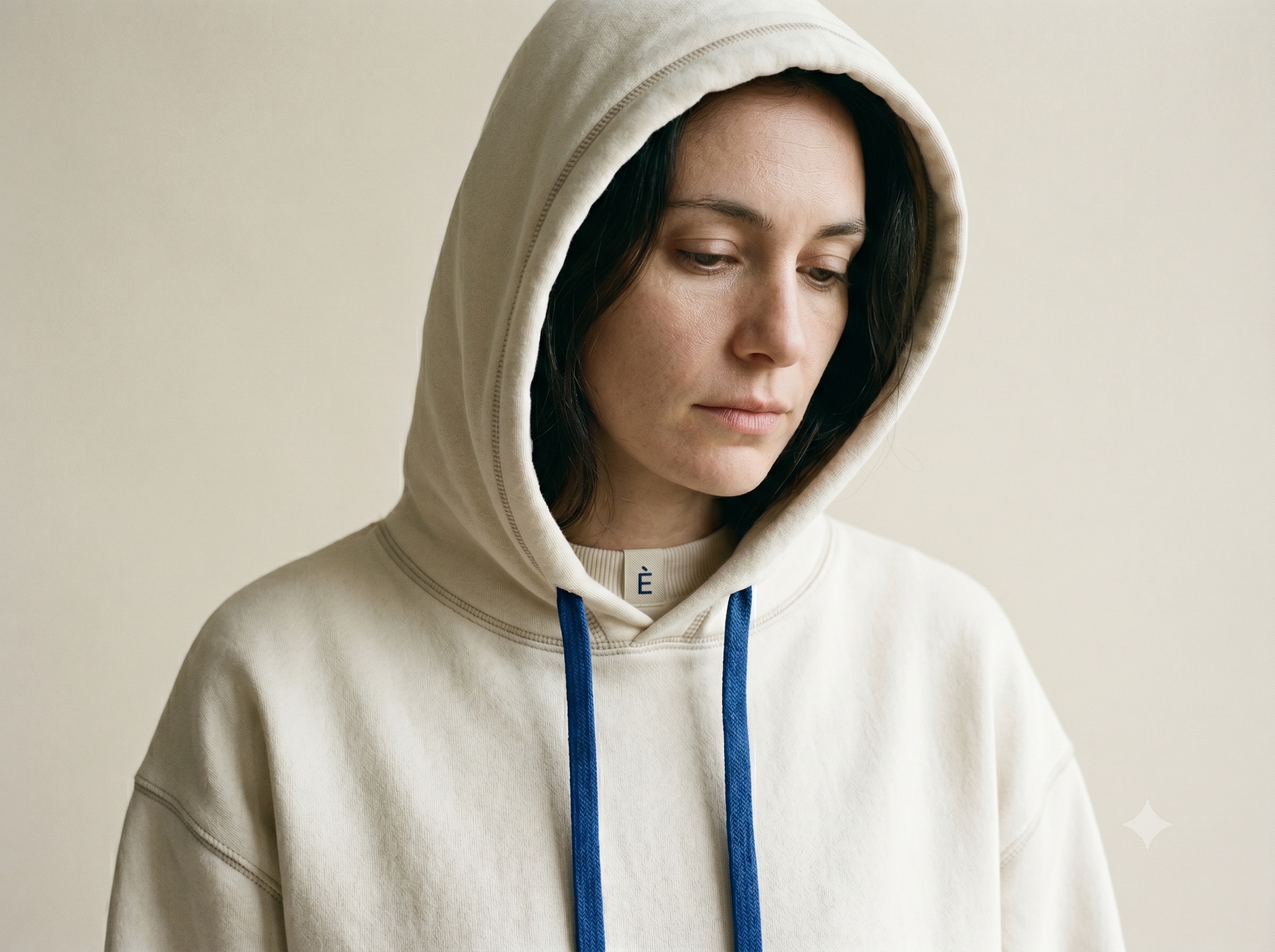 A calm, unsmiling person in a head-and-shoulders portrait wearing a relaxed ecru heavy-cotton hoodie with the deep hood drawn loosely up off the brow, both hood drawcords in ink-blue hanging down the chest, against a warm off-white studio backdrop in soft north light.