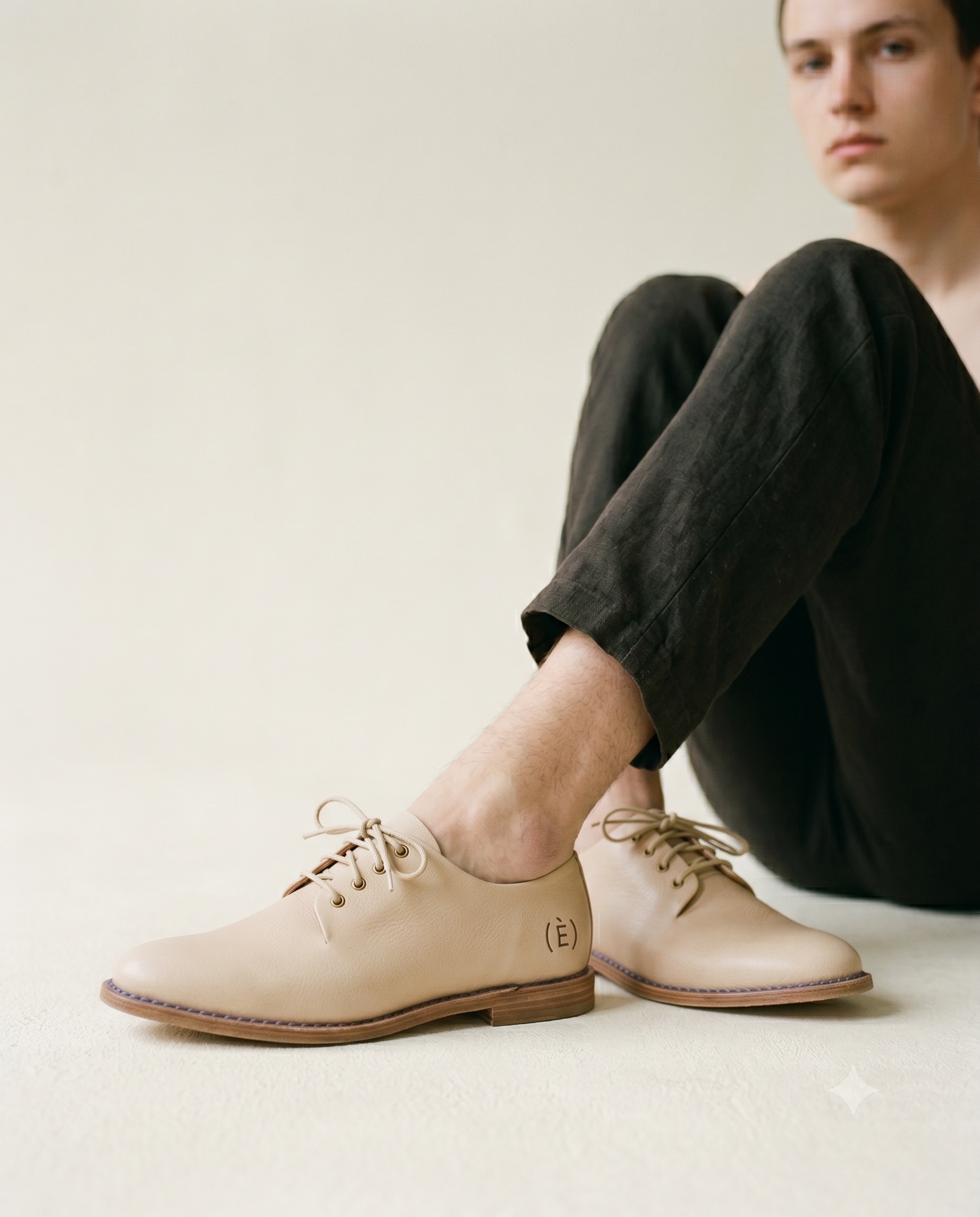 A seated model's feet at ground level wearing low ecru vegetable-tanned leather derby shoes on a continuous undyed-ecru studio floor, the upper cut as one unbroken line on a slim leather sole, lit by soft north-light; a single ink-blue welt thread runs between upper and sole and a small (È) grave seal is blind-embossed on the heel.