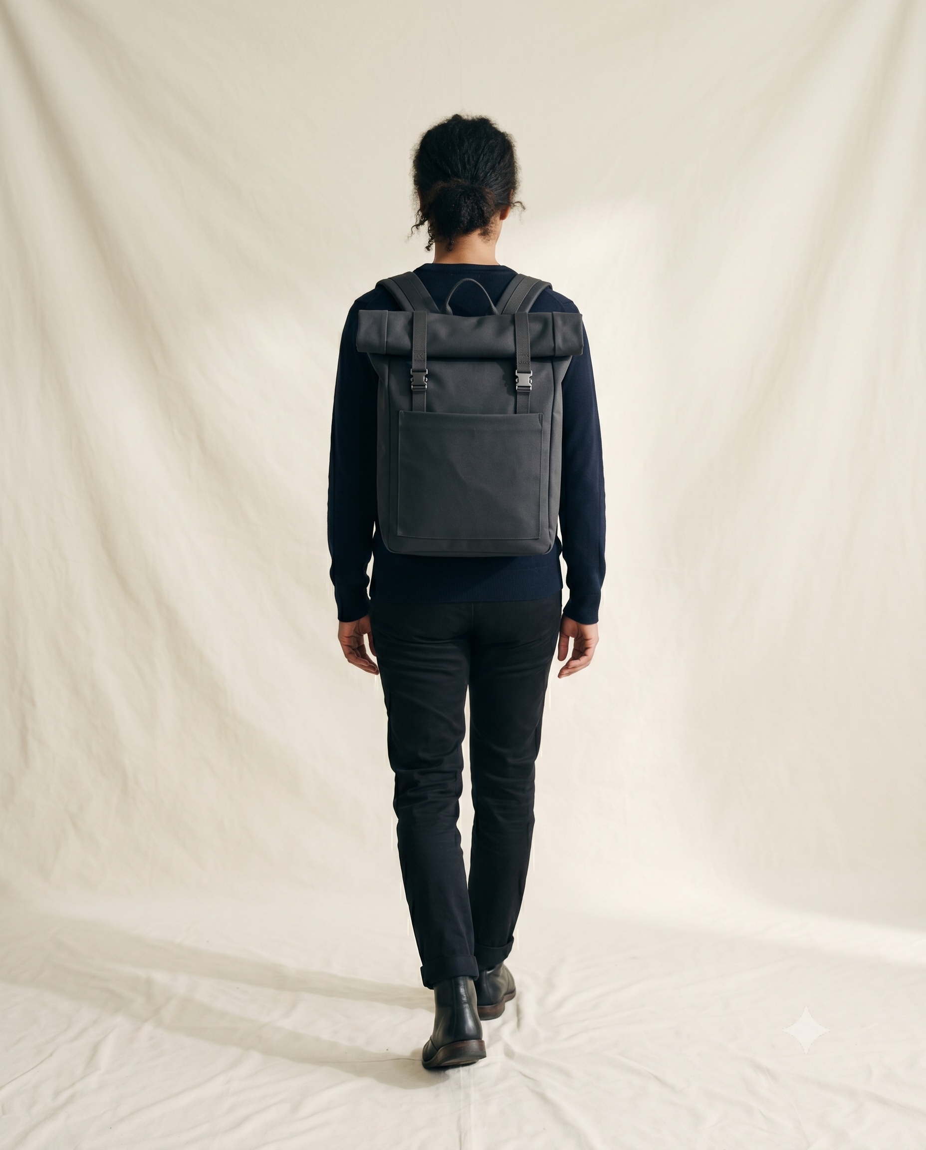 A person seen from directly behind walking slowly away from camera, wearing a squared, rigid deep-ink waterproof backpack with a welded lid, two compression catches and one bright ink-blue taped seam, against a plain warm ecru studio background in soft north-light.